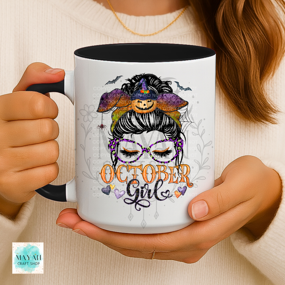 A person holds a white ceramic mug with a black interior and handle. The mug features a Halloween-themed illustration of a witch with purple glasses, a pumpkin hat, and the text 'OCTOBER Girl' in orange and purple.