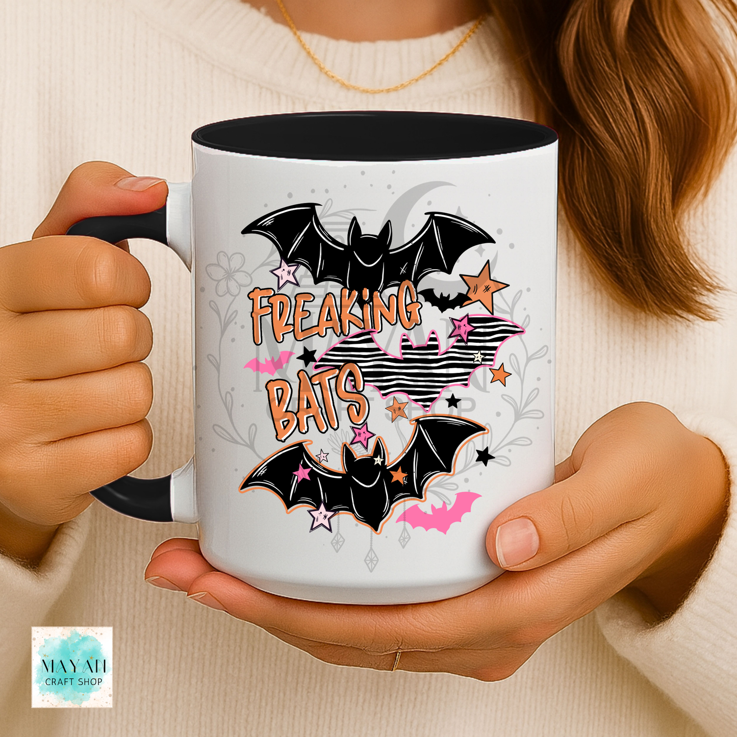 A person holds a white ceramic mug with a black handle and interior. The mug features a Halloween-themed graphic with two black bats, pink stars, and the words 'FREAKING BATS' in orange and pink.