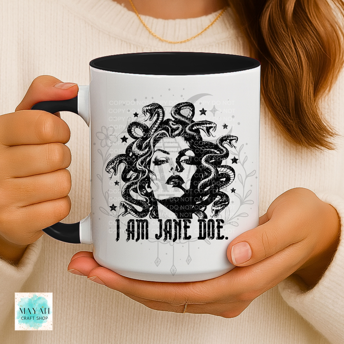 A person holds a white ceramic mug with a black handle and black interior. The mug features a black-and-gray illustrated portrait of Medusa with the text 'I AM JANE DOE.