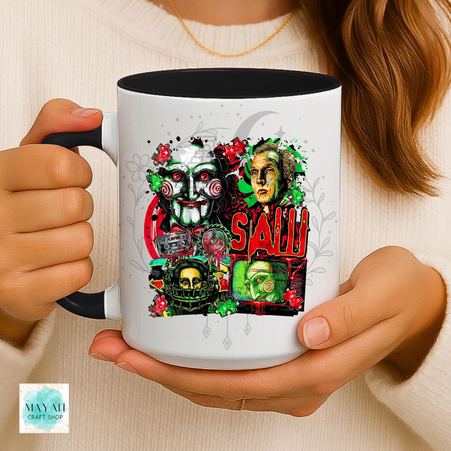A person holds a white ceramic mug with a black interior and handle. The mug features a colorful graphic collage of horror-themed faces, skulls, roses, and the a text in red.