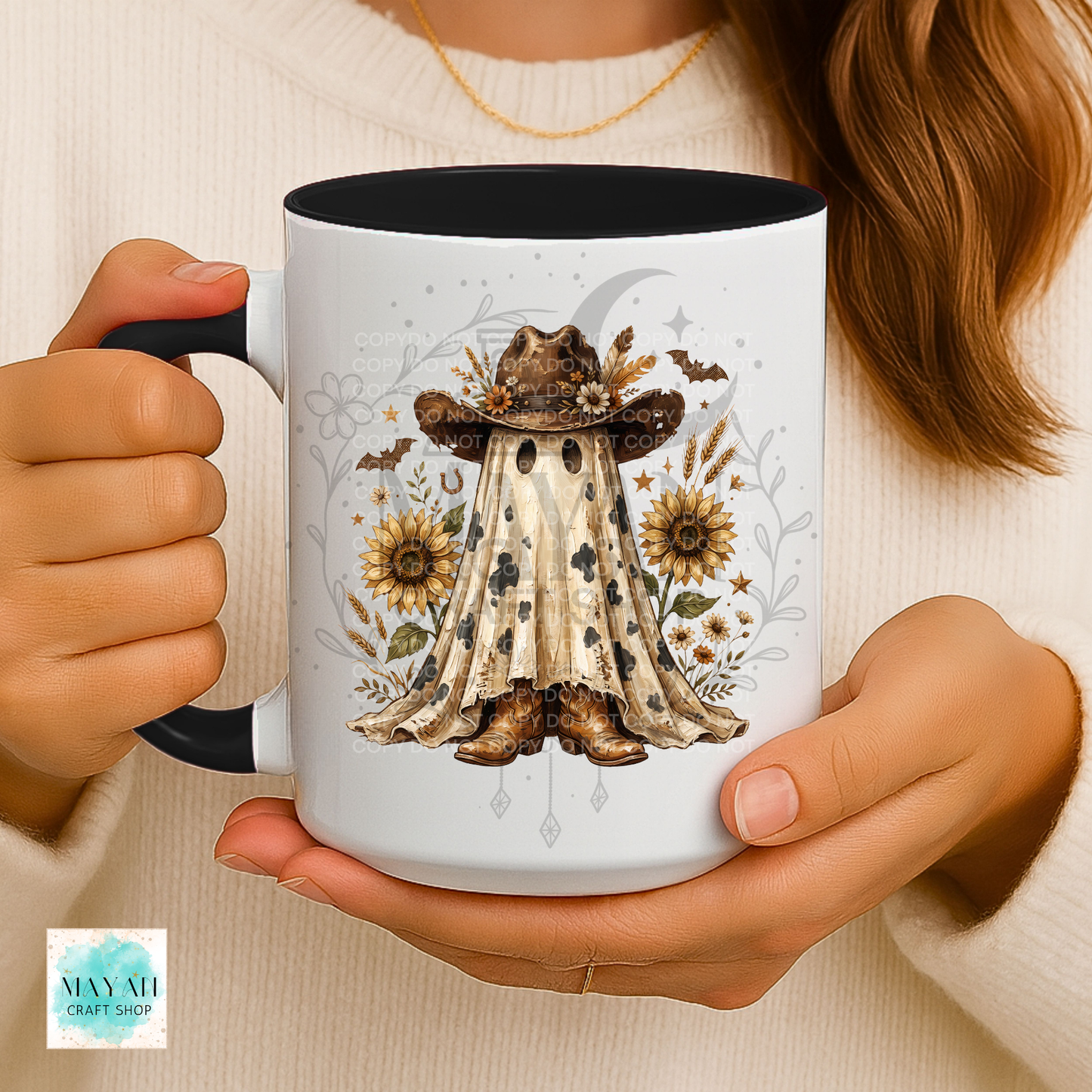 A person holds a white ceramic mug with a black interior and handle. The mug features a western-themed illustration of a ghostly figure in a cowboy hat surrounded by sunflowers, horseshoes, and a crescent moon.