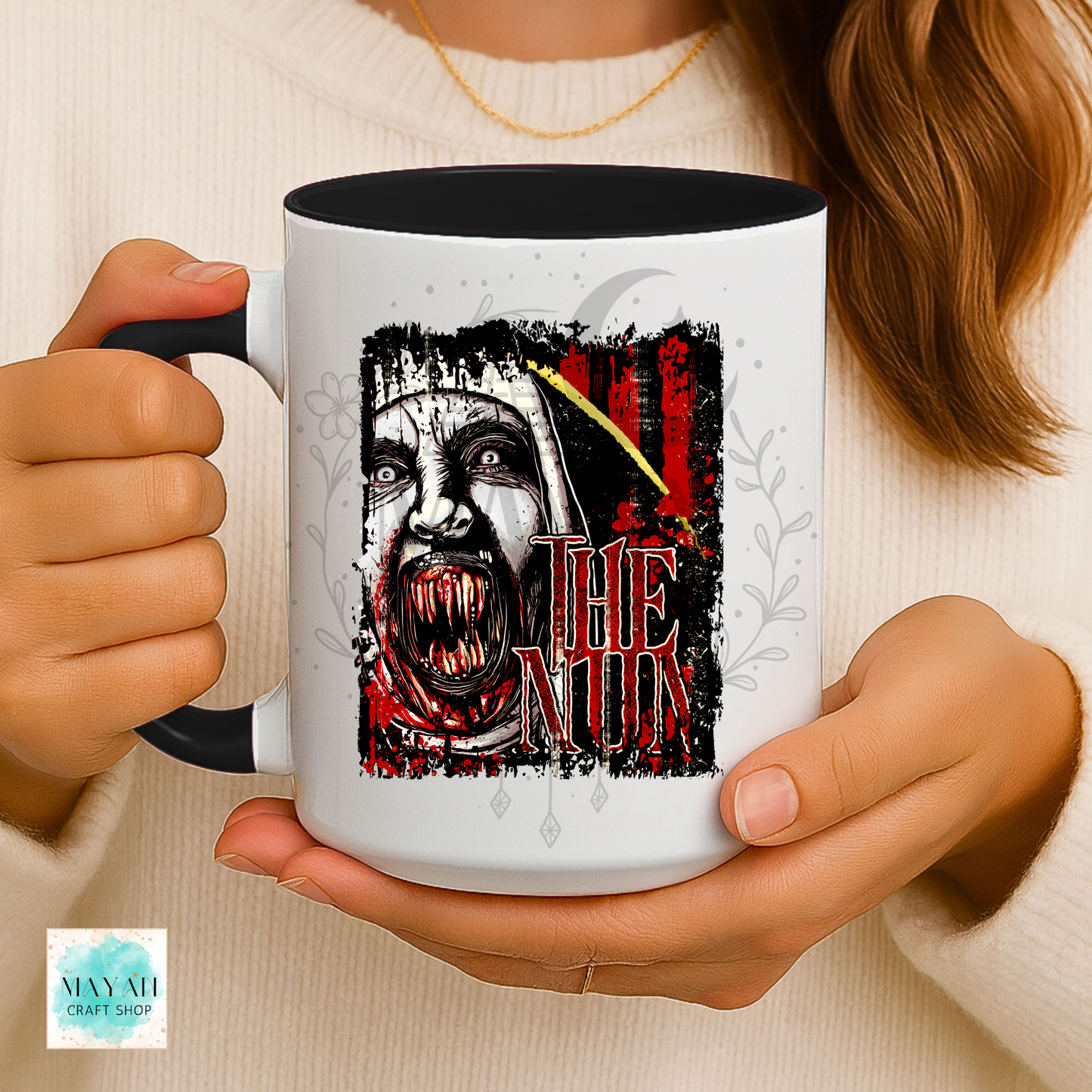 A person holds a white ceramic mug with a black interior and handle. The mug features a graphic print of a creepy nun face with a text in red, with a yellow crescent moon above.