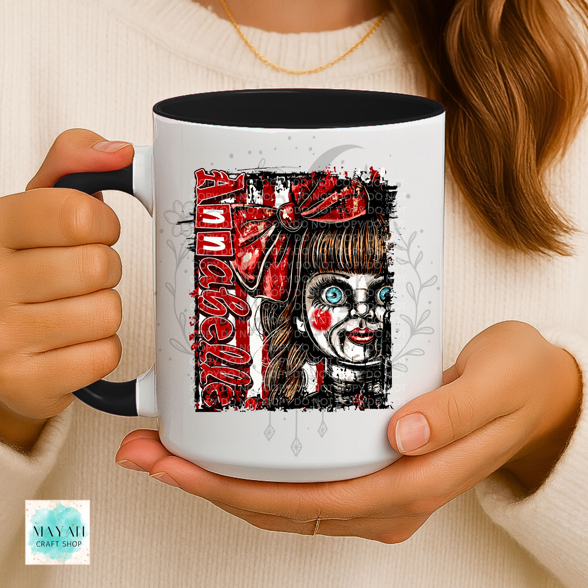 A person holds a white ceramic mug with a black handle and interior. The mug features a graphic print of a creepy doll-like character with red hair bows, red face paint, and a word in distressed red lettering.