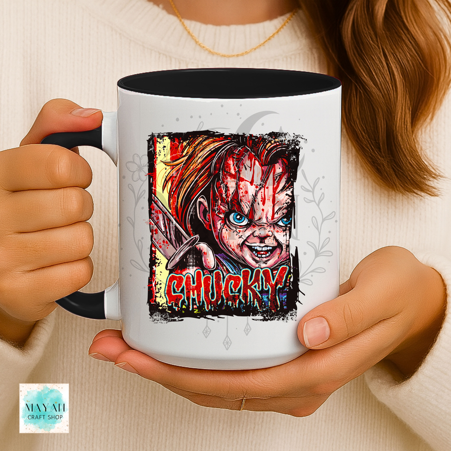 A person holds a white ceramic mug with a black handle and interior. The mug features a colorful graphic illustration of a doll with a word in red dripping letters.