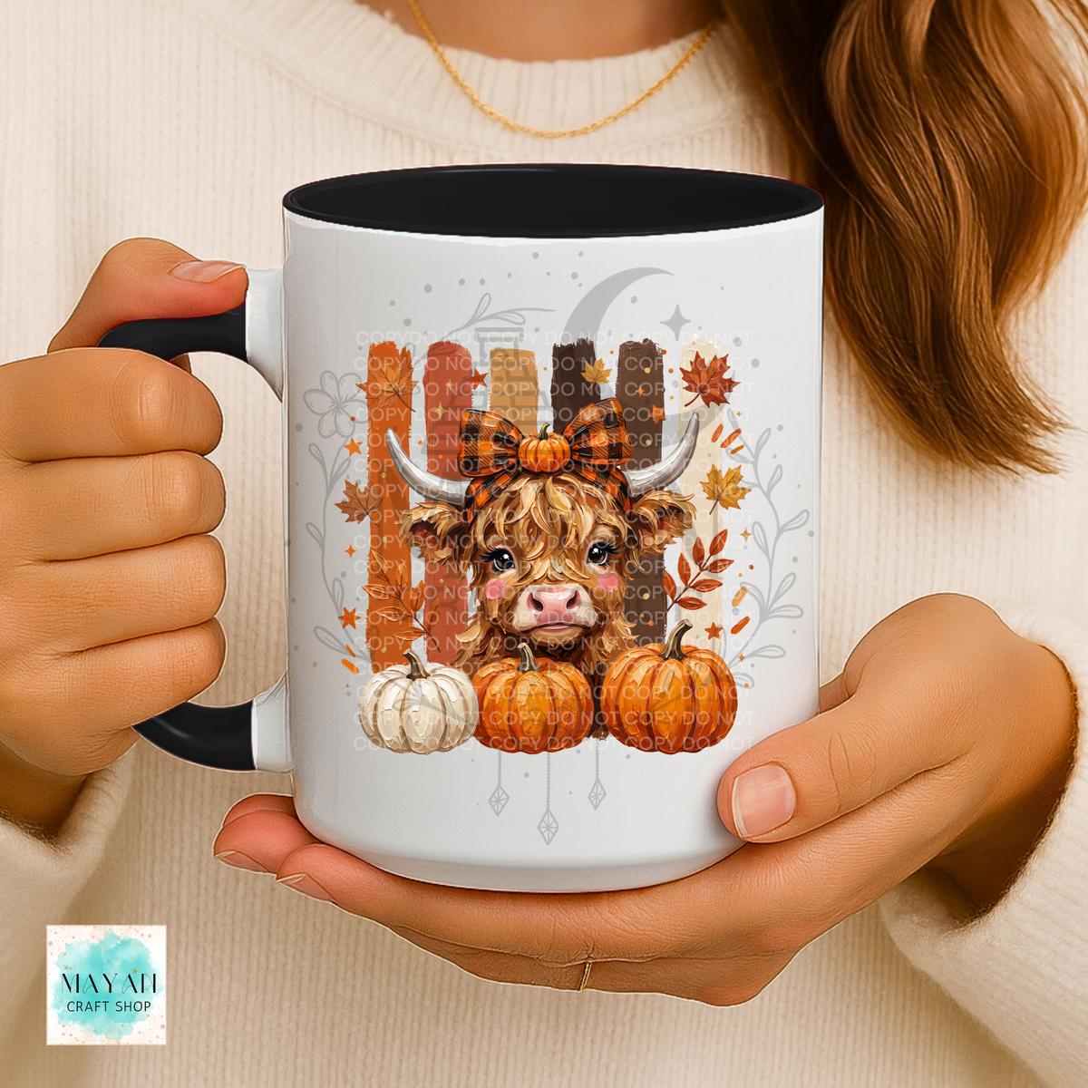 A person holds a white ceramic mug with a black interior and handle. The mug features a fall-themed illustration of a shaggy Highland cow with a plaid bow, pumpkins, autumn leaves, and a fence.