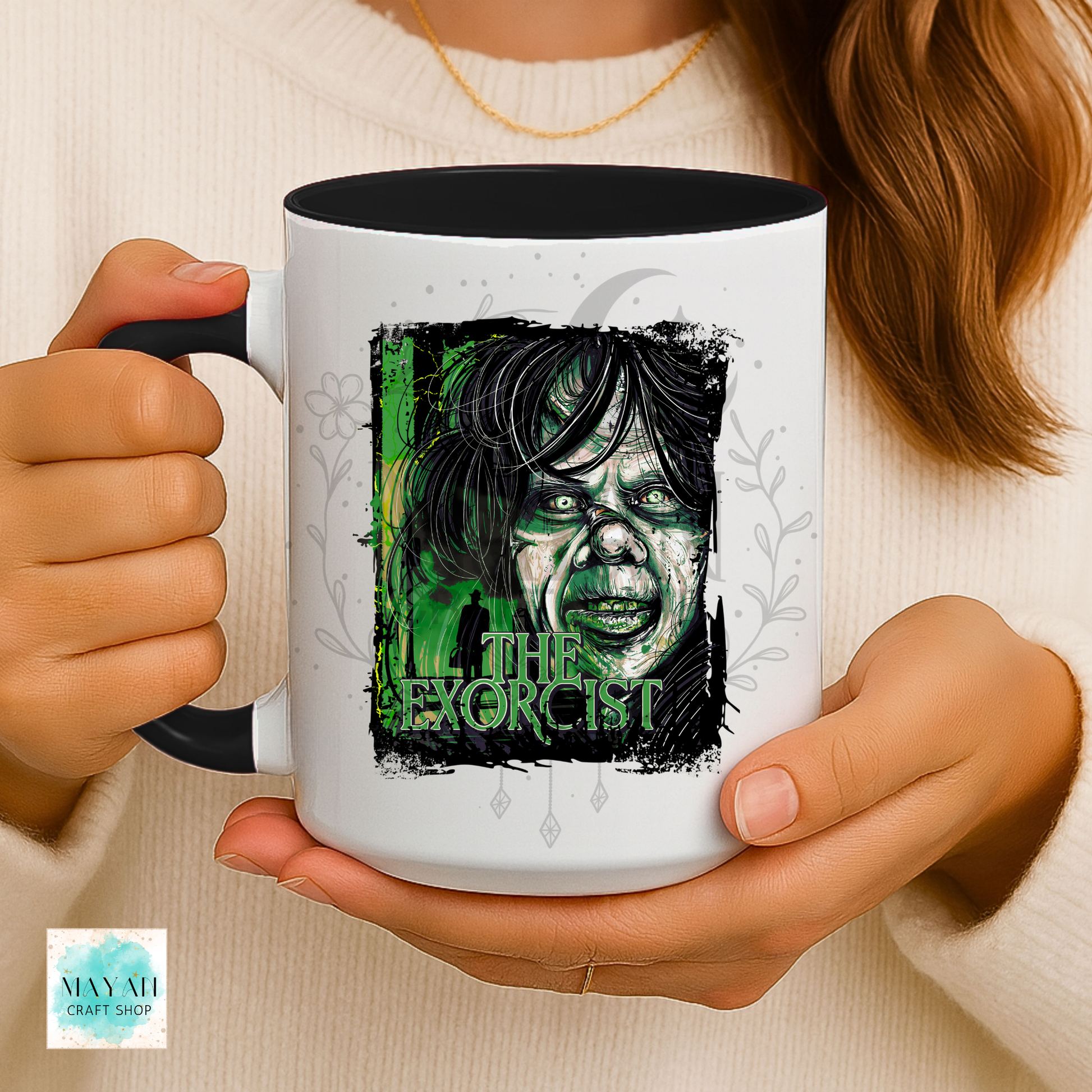 A person holds a white ceramic mug with a black interior and handle. The mug features a graphic print of a creepy zombie-like face with green and black tones and a text.