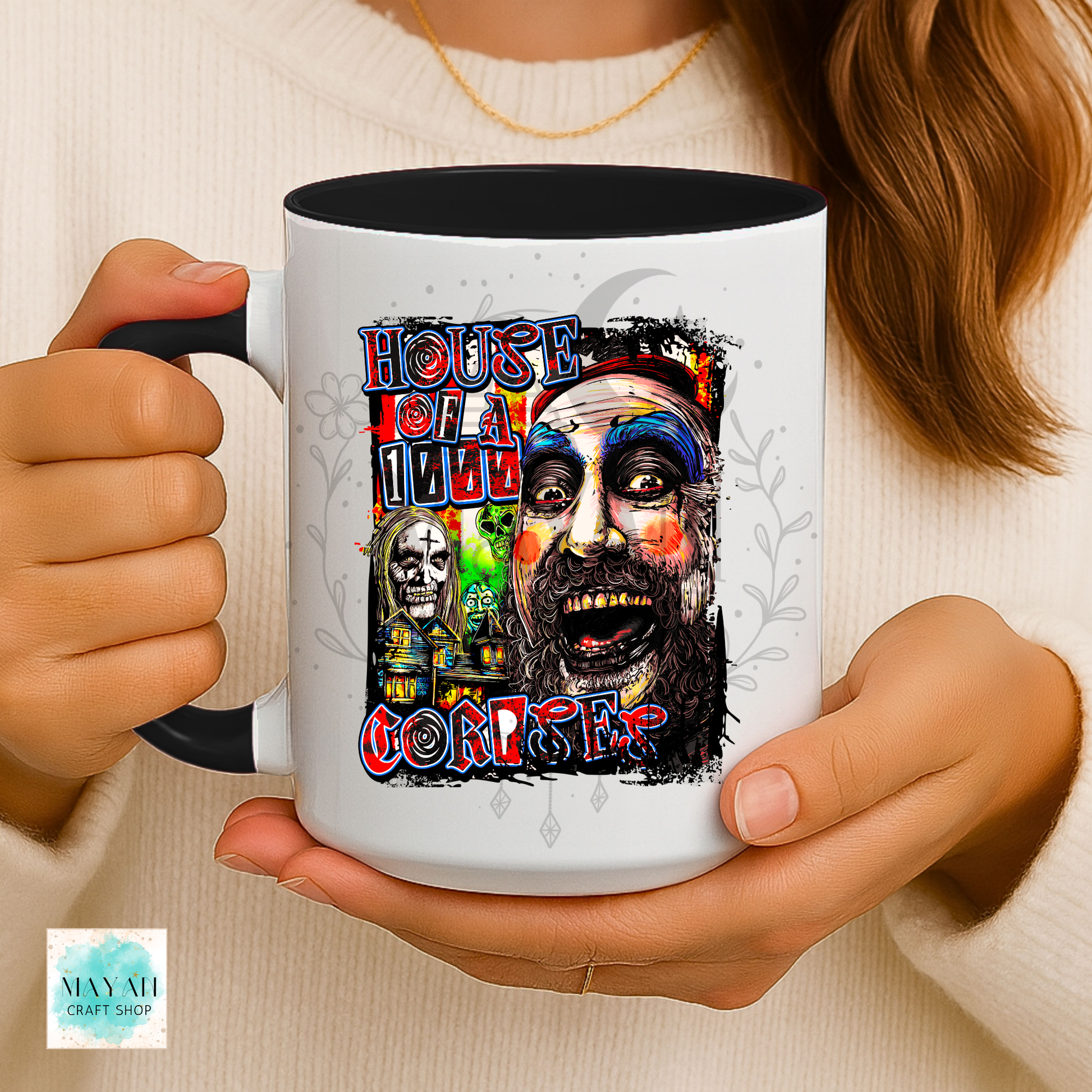 A person holds a white ceramic mug with a black handle and interior. The mug features a colorful graphic with a text and surreal illustrated faces.