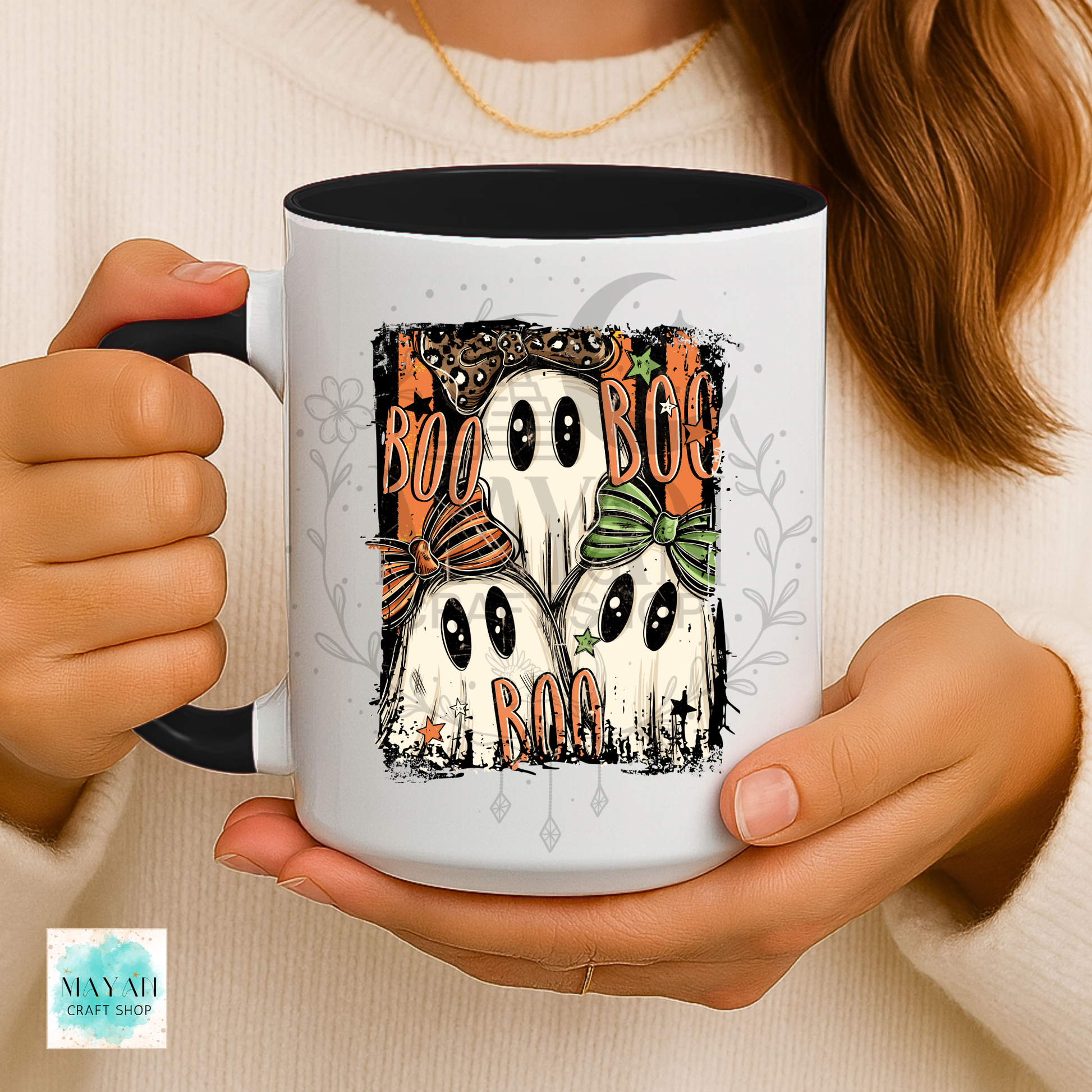 A person holds a white ceramic mug with a black handle and interior. The mug features a Halloween graphic of four cartoon ghosts with black eyes, orange bows, and the word 'BOO' in distressed orange lettering.
