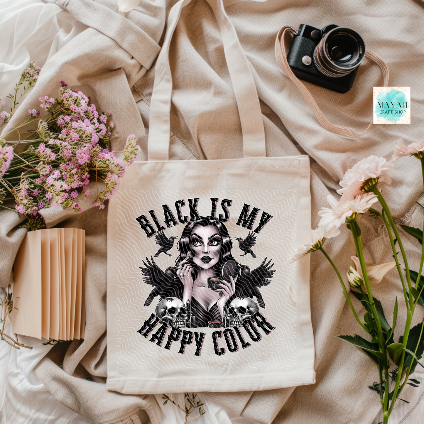 Beige tote bag with a graphic design and text on a soft fabric background with flowers and a camera.