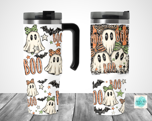 Two views of a stainless steel travel tumbler with a Halloween ghost pattern, shown on a gray wood plank background. The pattern features cute cartoon ghosts wearing green and orange bows, black bats, and the word 'BOO' in orange, with a black lid and handle.