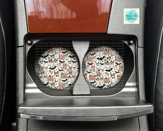 A pair of round car cup holders featuring a Halloween-themed pattern with ghosts, bats, pumpkins, and 'BOO' text, installed in a car's center console.