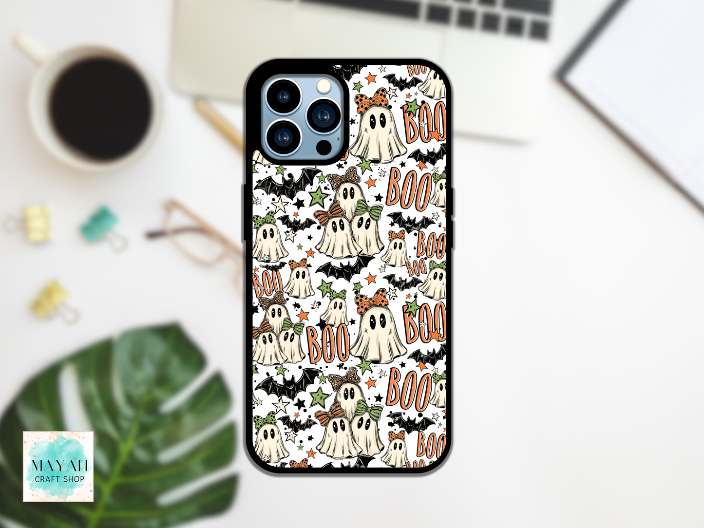 A phone case with a Halloween pattern featuring cartoon ghosts, bats, and 'BOO' text, shown on a phone in a styled desk flat-lay with a coffee cup, laptop, and green leaf.