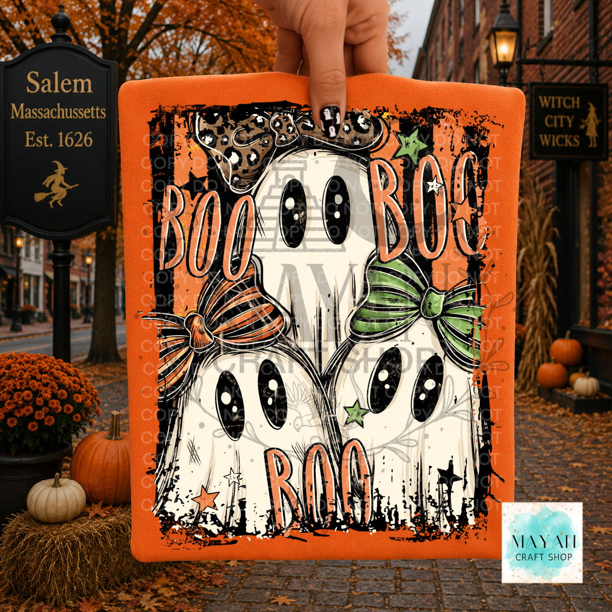 A hand holds a shirt featuring two cartoon ghosts with black eyes, orange bows, and the words “BOO BOO BOO.” The sign is photographed outdoors on a cobblestone street with pumpkins, autumn leaves, and storefront signs in the background.