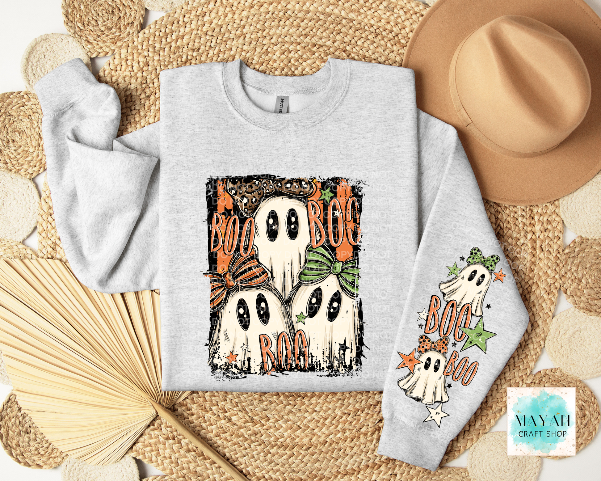 A light gray long-sleeve crewneck sweatshirt with a Halloween graphic print featuring four ghosts and the words 'BOO'. The sweatshirt is displayed in a flat-lay arrangement on a woven mat with a tan hat, a paper fan.