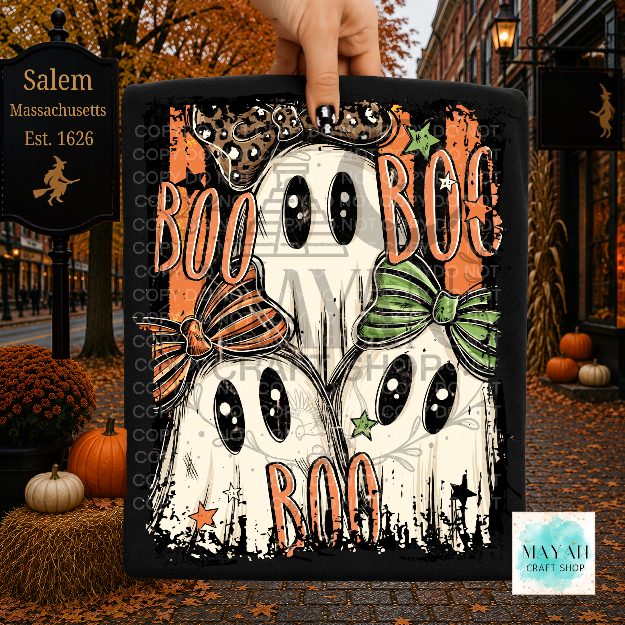 A hand holds a square black canvas print featuring a Halloween design of two white ghosts with black eyes, orange 'BOO' lettering, and bows in orange and green. The background depicts a fall street scene with pumpkins and a Salem.