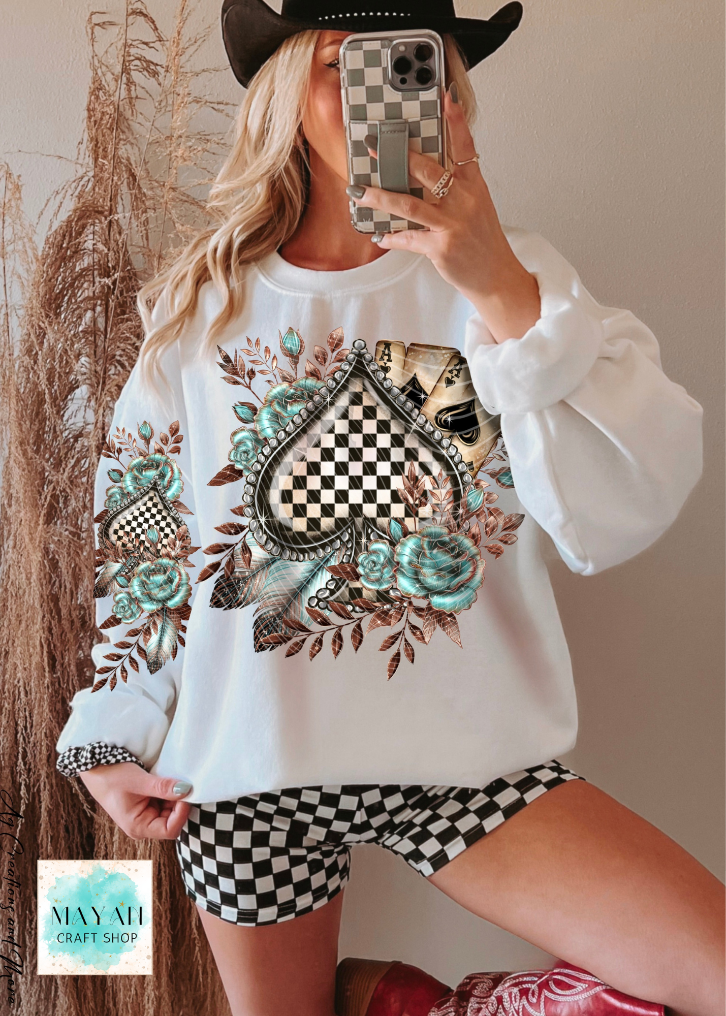 Person wearing a white sweatshirt with a decorative design and checkered shorts, taking a mirror selfie.