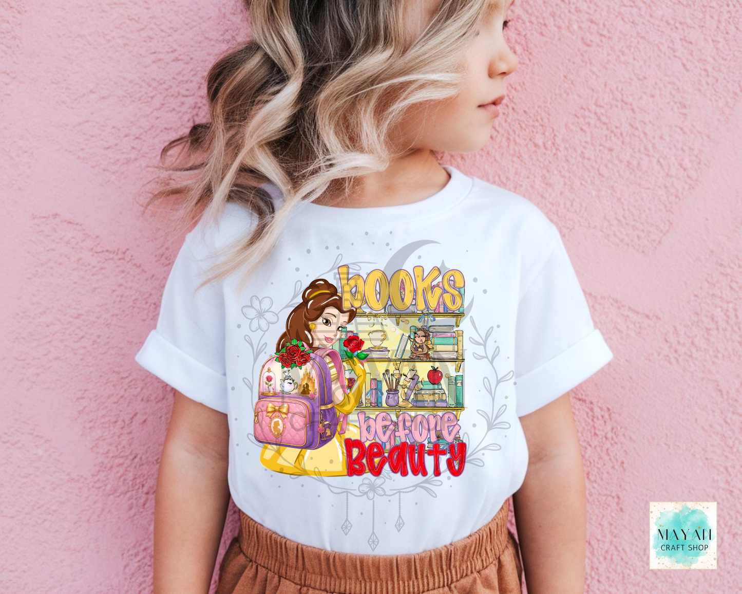 A child wearing a white graphic T-shirt with a colorful illustration  and the words 'BOOKS' before BEAUTY', posed against a pink textured wall.