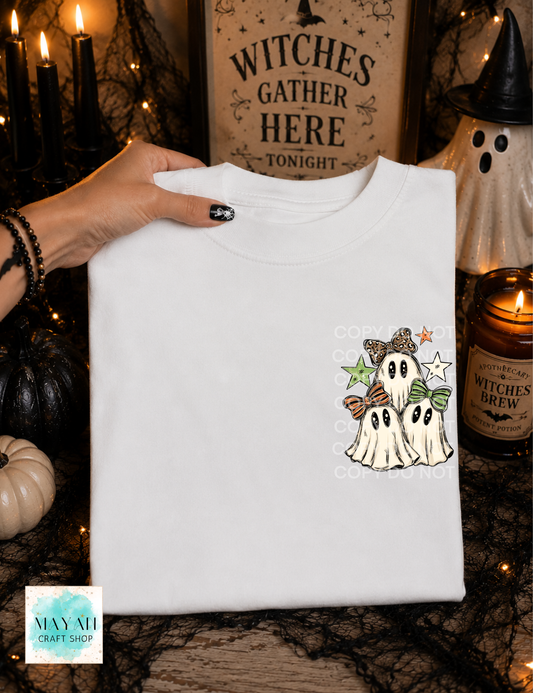 A folded white t-shirt with a cute cartoon ghost graphic, held by a hand in a spooky Halloween setting with candles, a witch hat, and a sign.