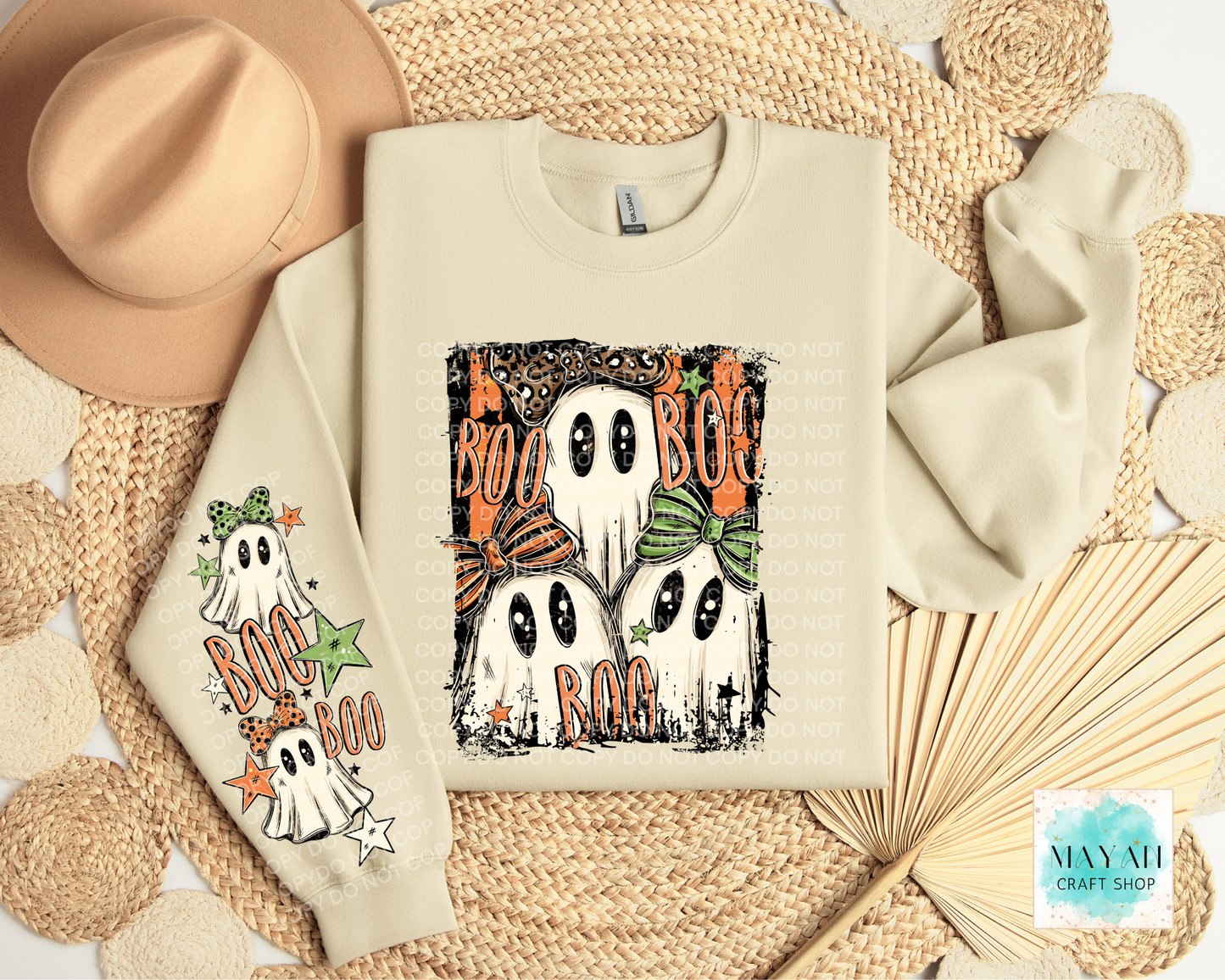 A cream-colored crewneck sweatshirt with Halloween graphics, displayed in a flat-lay arrangement with a beige hat, woven placemats, and a paper fan. The sweatshirt features a graphic of three ghosts with orange and green bows and the words 'BOO' on the front, and a matching graphic on the left sleeve.