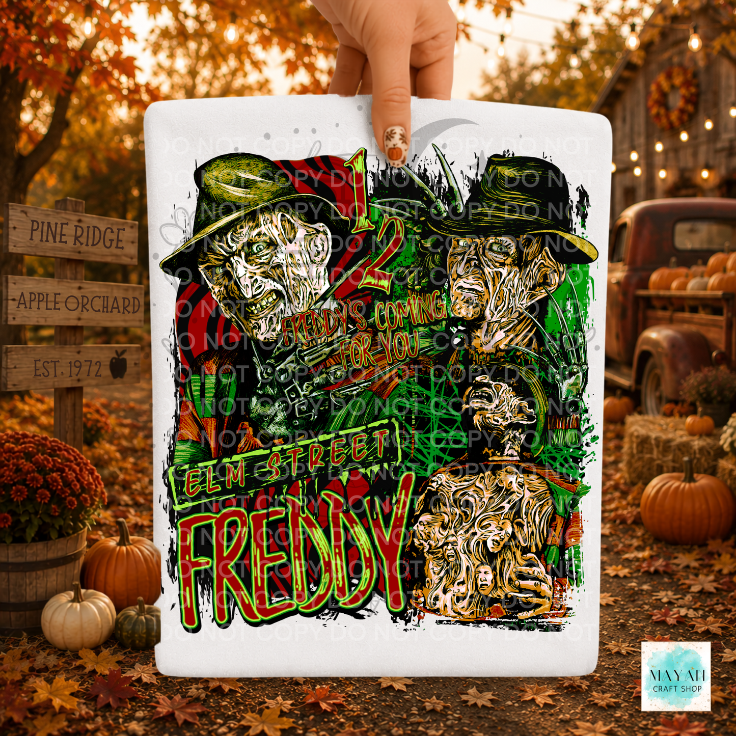 A hand holds a white shirt featuring a colorful graphic of two stylized Faces with a text. The background is a blurred autumn outdoor scene with pumpkins, fall leaves, and rustic decor.