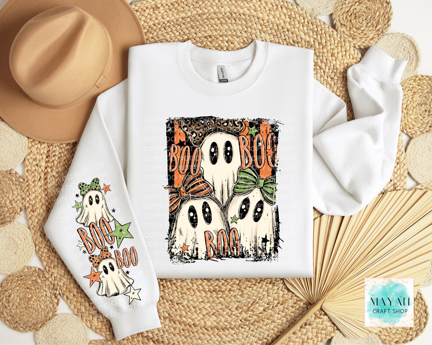 A folded white sweatshirt with Halloween graphics, displayed on a woven beige mat with a tan hat, paper fans, and decorative circles. The sweatshirt features a large front graphic of three ghosts with orange bows and the word 'BOO', plus a smaller matching graphic on the sleeve.