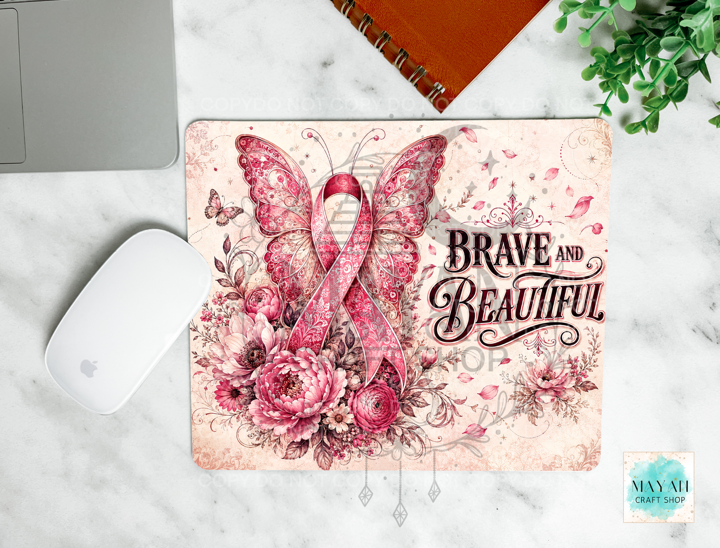 A mouse pad with a pink and red floral design featuring a butterfly, roses, and the words 'BRAVE AND BEAUTIFUL,' shown on a white marble surface with a mouse, laptop edge, notebook, and plant nearby.