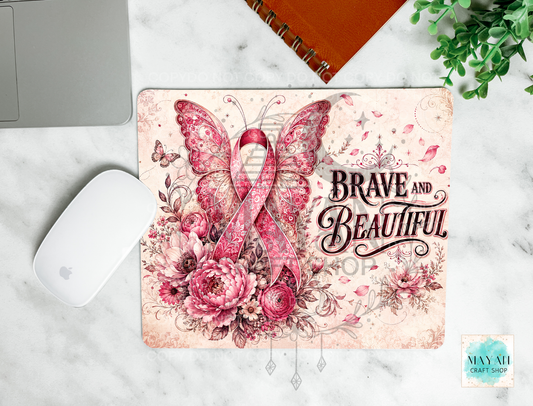 A mouse pad with a pink and red floral design featuring a butterfly, roses, and the words 'BRAVE AND BEAUTIFUL,' shown on a white marble surface with a mouse, laptop edge, notebook, and plant nearby.