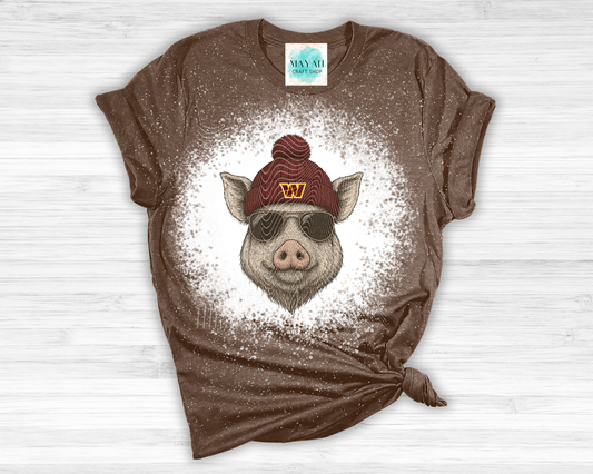 Brown t-shirt with a graphic of a pig wearing a red beanie and sunglasses on a white wooden surface.