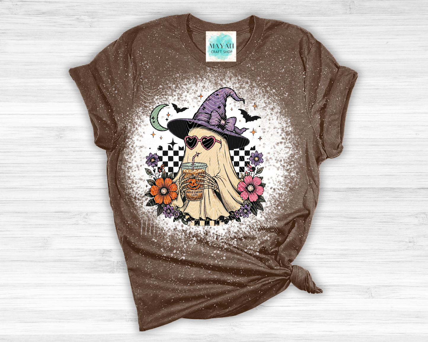 Brown t-shirt with a ghost wearing a witch hat and sunglasses graphic design on a white wooden surface.