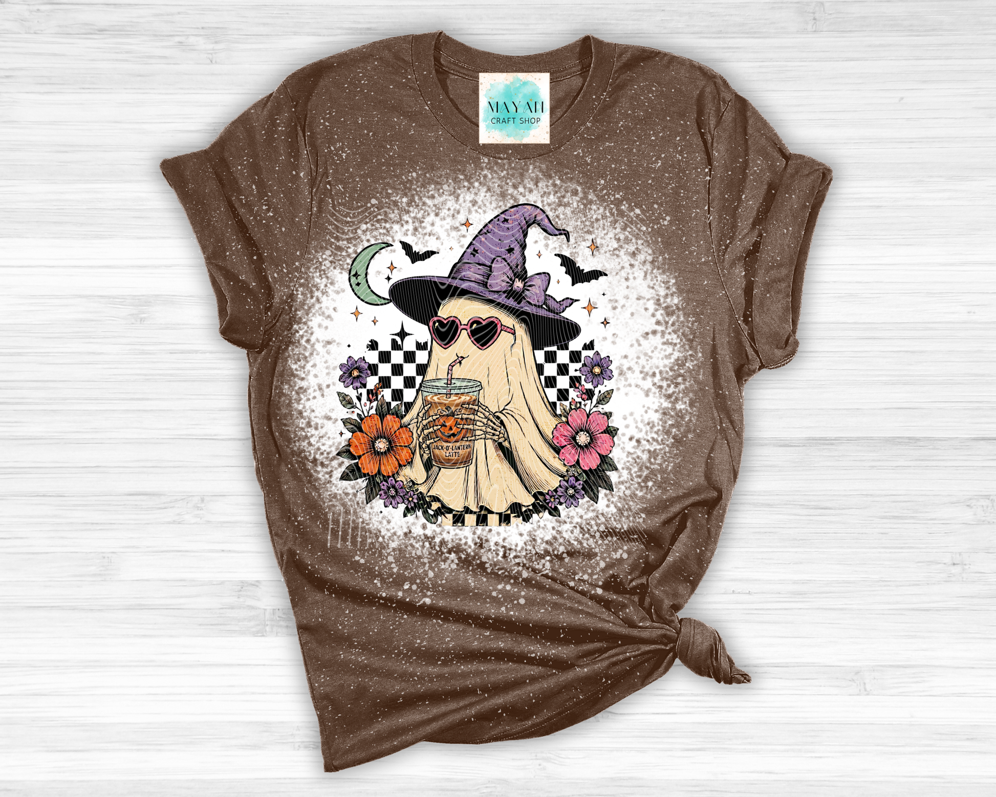 Brown t-shirt with a ghost wearing a witch hat and sunglasses graphic design on a white wooden surface.