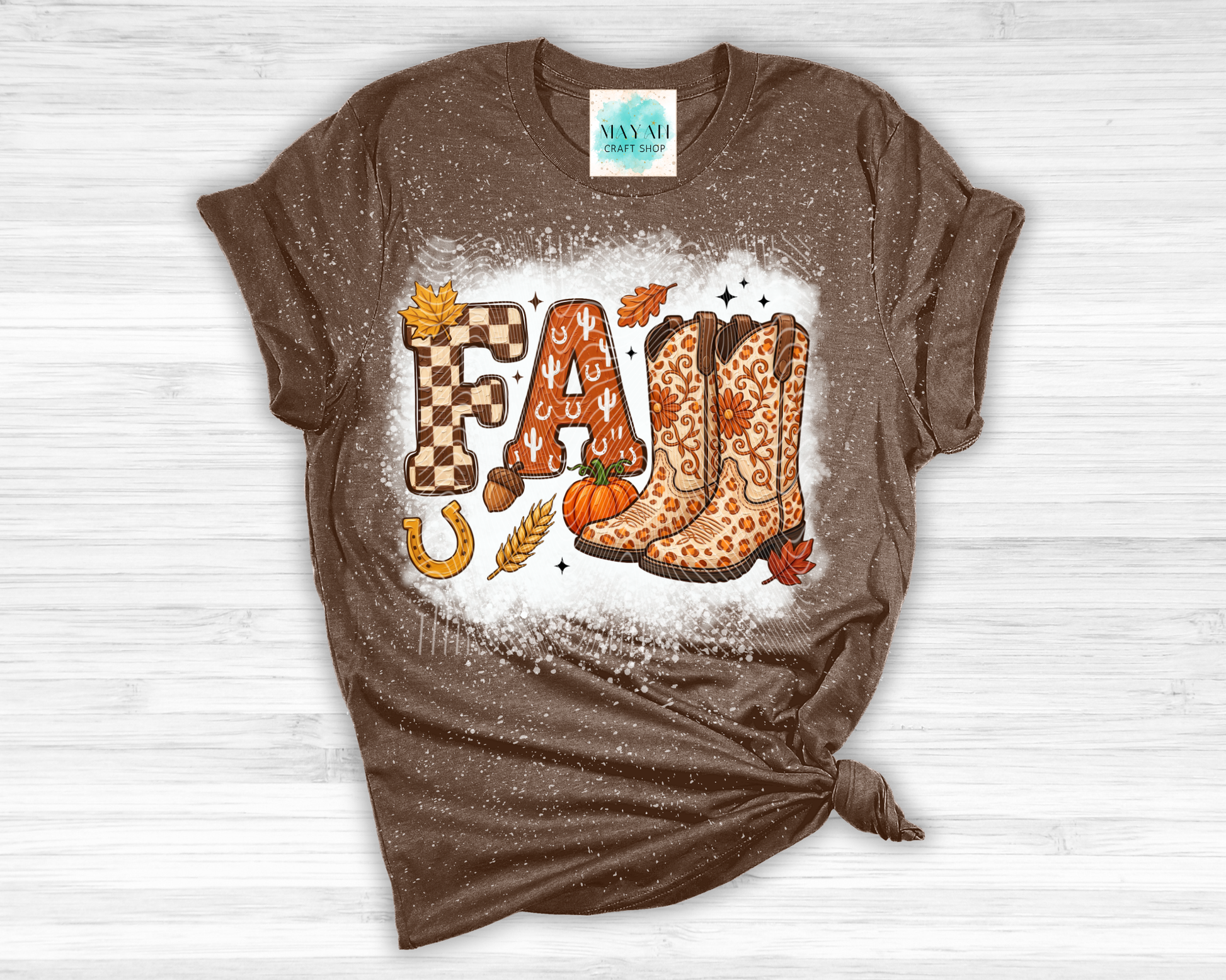 A brown short-sleeve t-shirt with a distressed texture, featuring a 'FALL' graphic with cowboy boots, autumn leaves, and a pumpkin on a white splattered background. The shirt is styled with a knot at the bottom right and is laid flat on a white wood background.