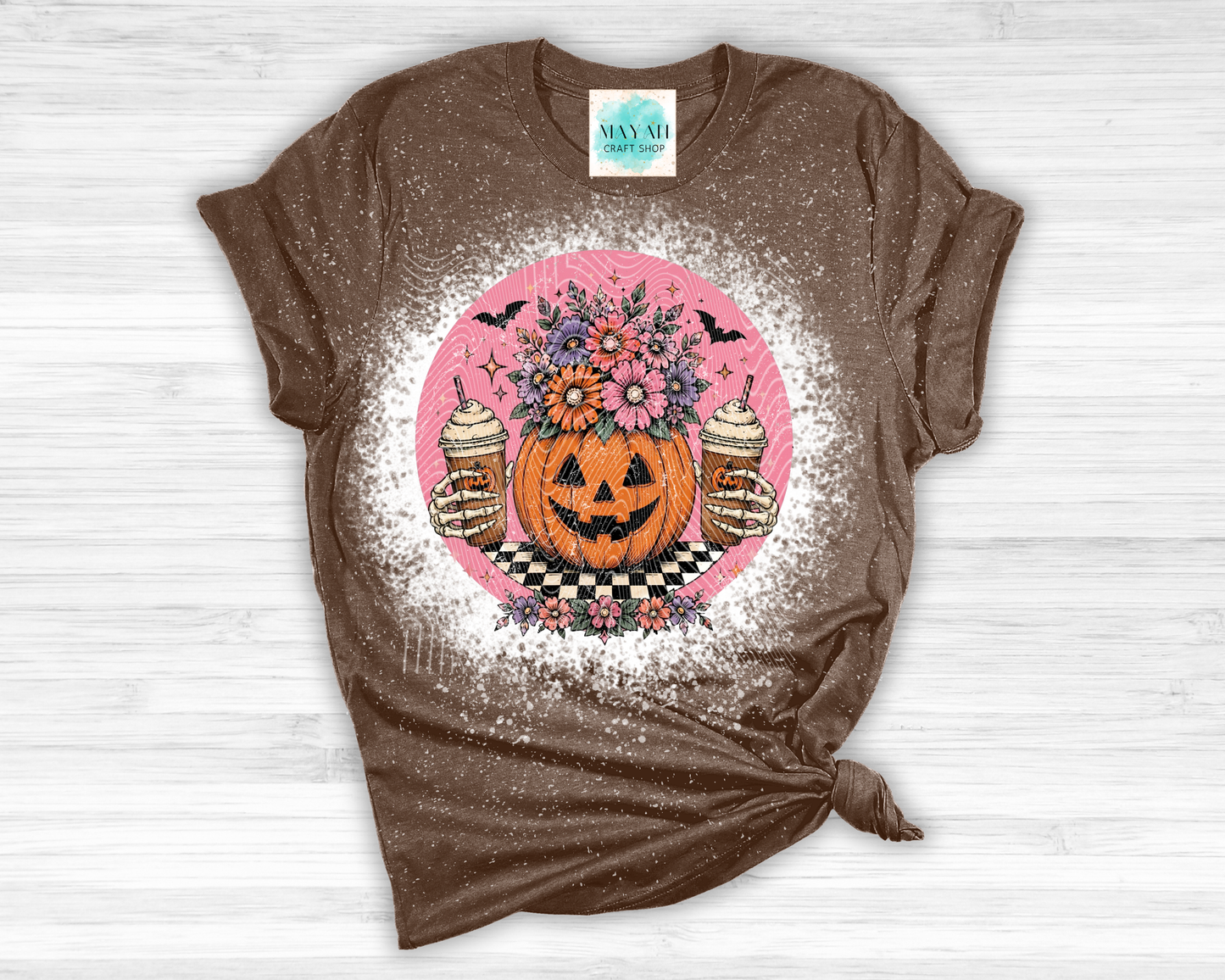 Brown t-shirt with a Halloween-themed design featuring a pumpkin, flowers, and coffee on a white wooden background.