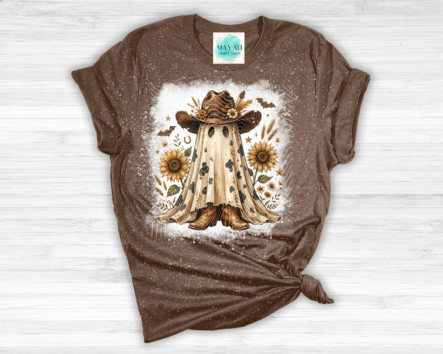 A brown short-sleeve t-shirt with a distressed texture, featuring a central graphic of a ghostly figure in a cowboy hat surrounded by sunflowers, laid flat on a white wood background.
