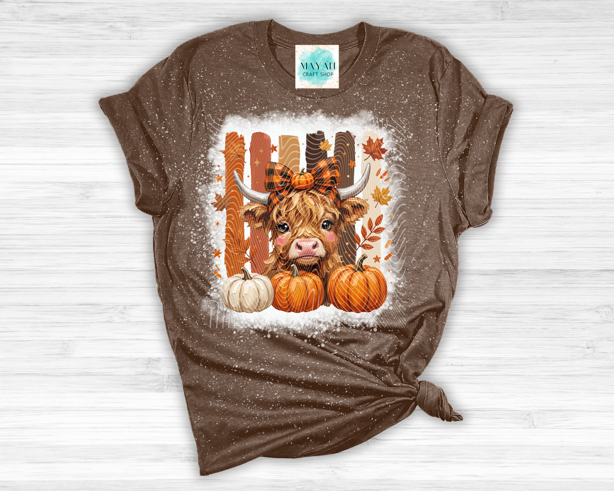 A brown short-sleeve t-shirt with a knotted hem, featuring a central autumn graphic of a Highland cow, pumpkins, and a fence decorated with fall leaves. The shirt is displayed flat on a light gray wood-plank background.