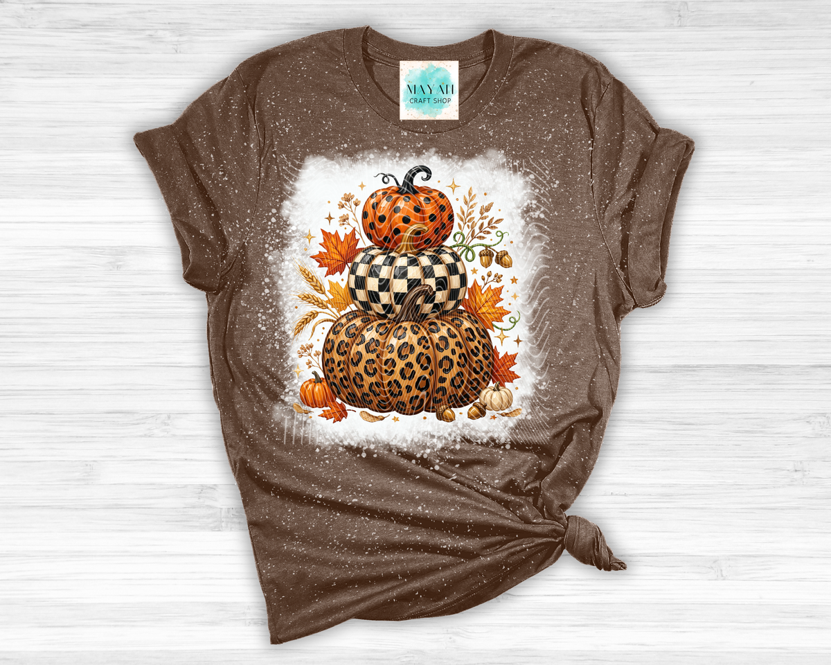 A brown short-sleeve t-shirt with a distressed, faded texture, featuring a central graphic of stacked pumpkins in leopard and checkered patterns surrounded by autumn leaves. The shirt is styled flat on a white wood background with a knot at the bottom hem.