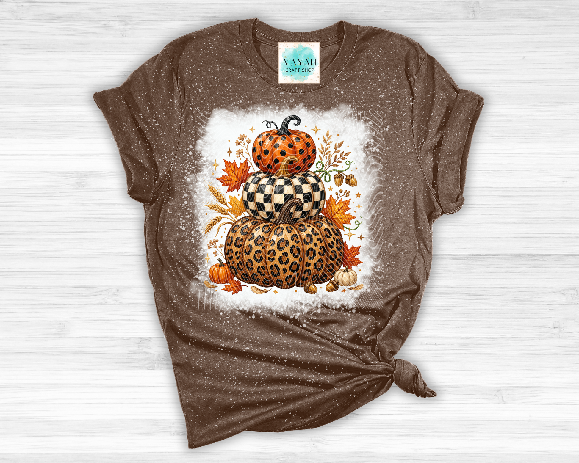 A brown short-sleeve t-shirt with a distressed, faded texture, featuring a central graphic of stacked pumpkins in leopard and checkered patterns surrounded by autumn leaves. The shirt is styled flat on a white wood background with a knot at the bottom hem.