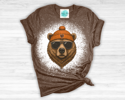 Brown t-shirt with a bear graphic wearing sunglasses and an orange beanie on a white wooden surface.