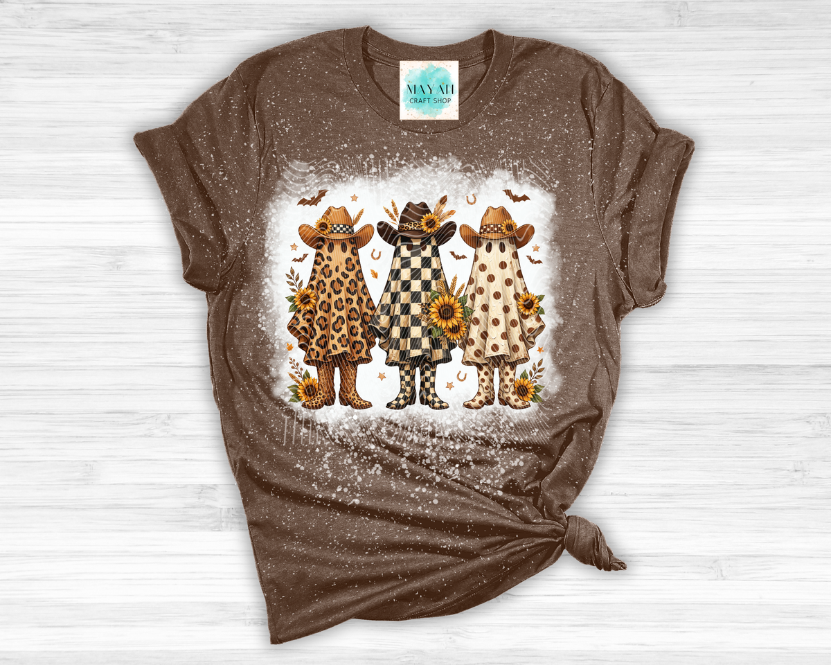 A brown short-sleeve bleached t-shirt featuring a graphic of three western-style ghosts wearing cowboy hats and boots. The shirt is styled with a knot at the bottom hem and is laid flat on a white wood background.