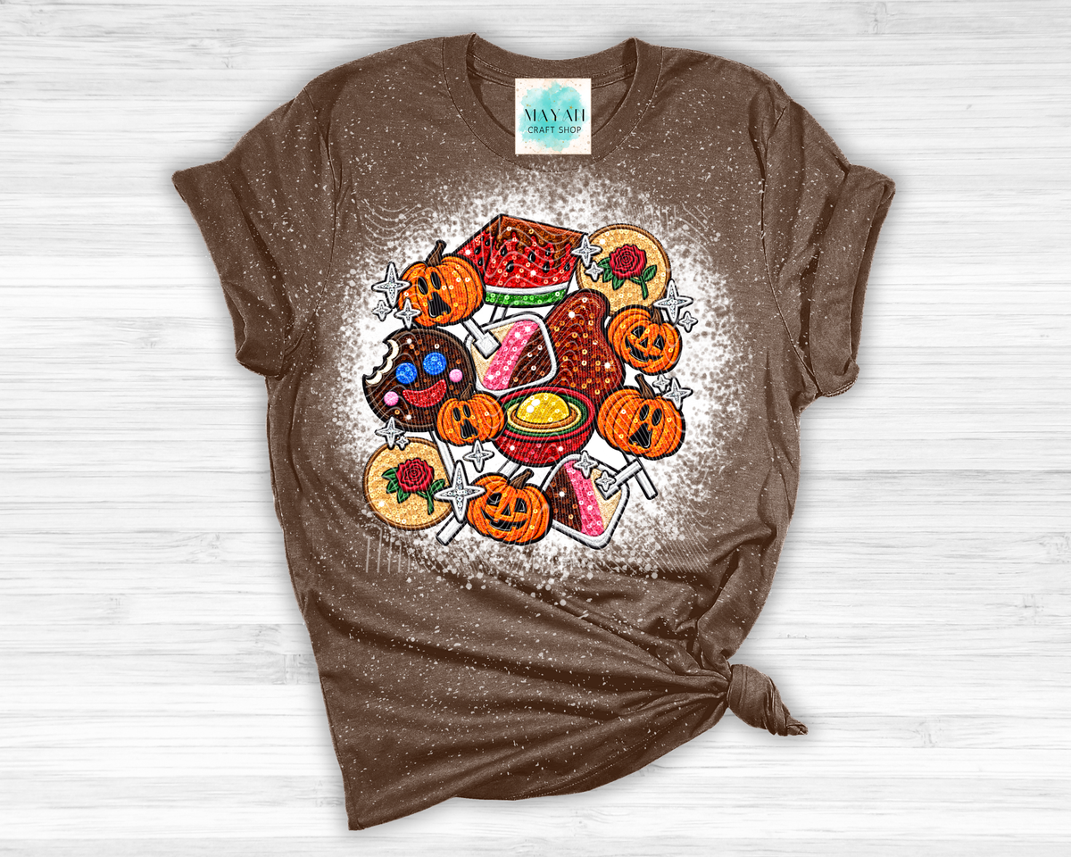 A brown short-sleeve t-shirt with a Halloween-themed graphic print of cute, colorful treats like pumpkins, donuts, and candy corn, styled on a white wood background.