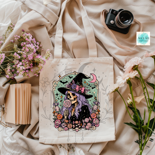 Tote bag with a colorful witch design on a soft fabric background with flowers and a camera.