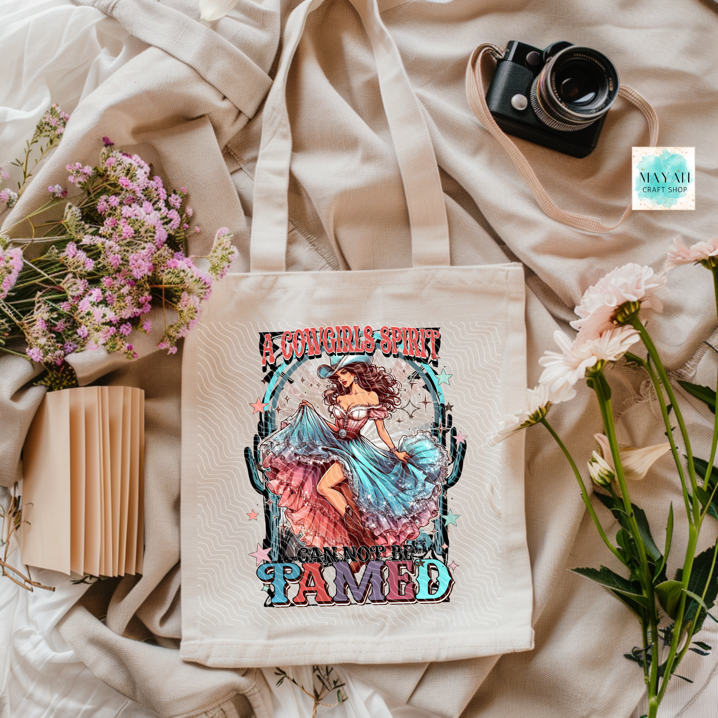 Tote bag with artistic design featuring a woman and text, surrounded by flowers and a camera on a soft fabric background.