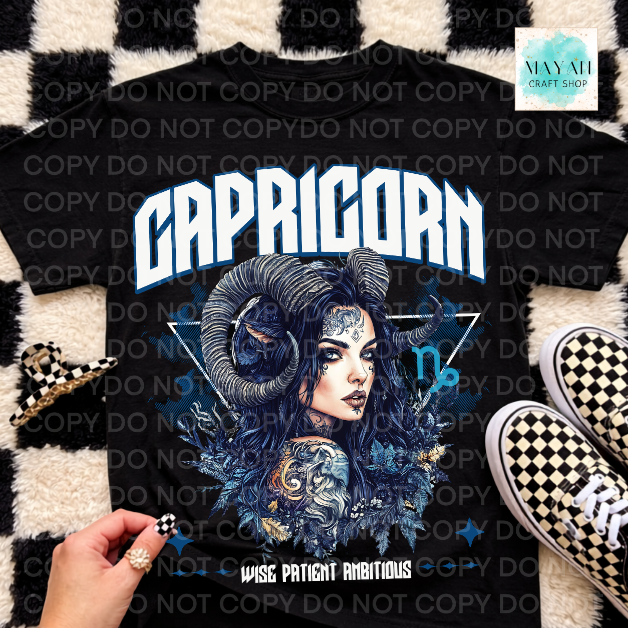A black graphic t-shirt featuring a detailed Capricorn illustration with a ram's head, a woman's face, and the word 'CAPRICORN' in bold white letters. The shirt is displayed in a flat-lay arrangement on a black and white checkered rug, next to a hand, a small checkered accessory, and black-and-white checkered sneakers.