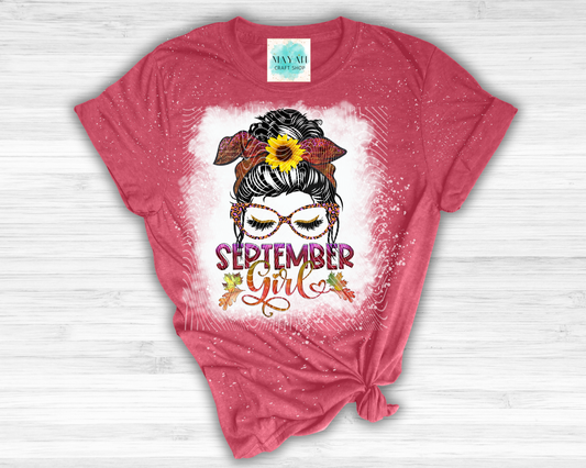 A heathered cardinal red graphic t-shirt with a 'September Girl' autumn theme, featuring a sunflower, fall leaves, and glasses on a white splattered background, styled flat on a light wood surface.