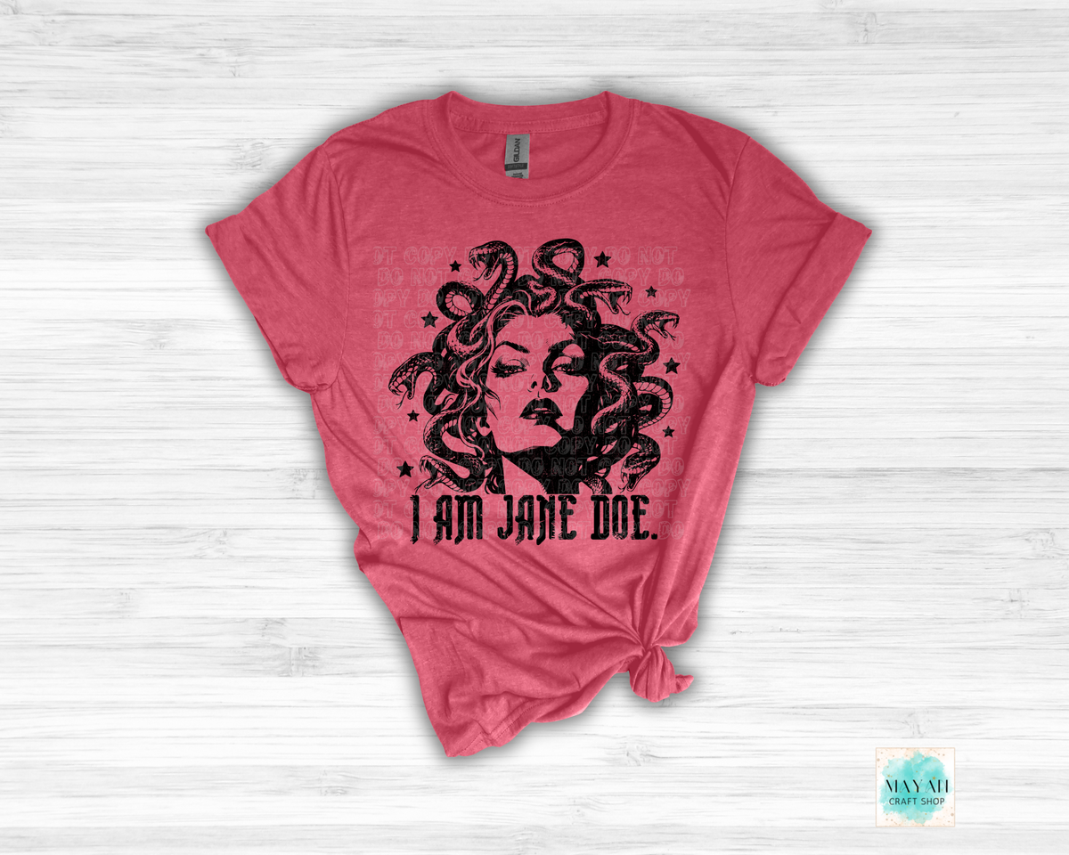 A pink short-sleeve t-shirt with a black graphic of Medusa and the text 'I AM JANE DOE.' printed on the front, styled flat on a white wood background.
