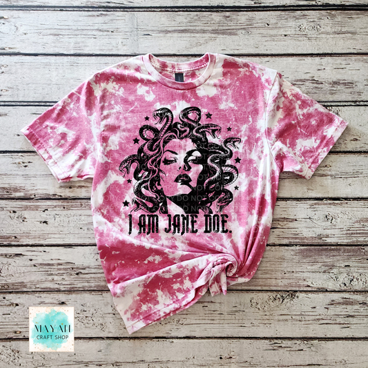 A heather cardinal red bleached graphic t-shirt featuring a black illustration of Medusa and the text 'I AM JANE DOE.' laid flat on a rustic wooden plank background.
