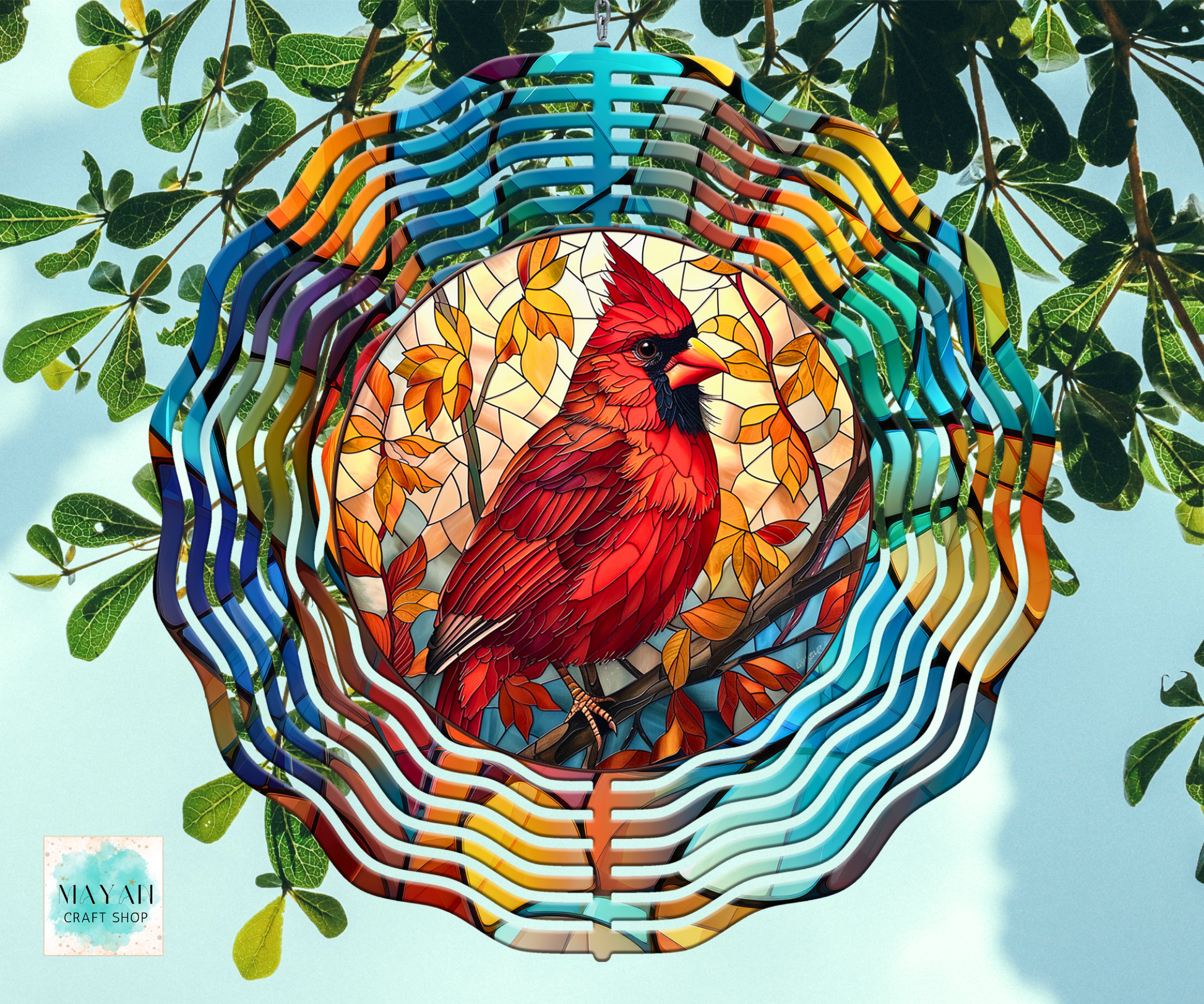 A colorful stained-glass-style wind spinner featuring a red cardinal perched on autumn branches, surrounded by wavy multicolored bars and hanging from green branches against a pale blue sky. 
