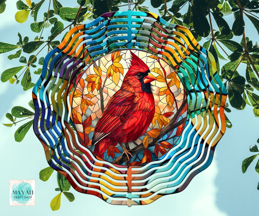 A colorful stained-glass-style wind spinner featuring a red cardinal perched on autumn branches, surrounded by wavy multicolored bars and hanging from green branches against a pale blue sky. 