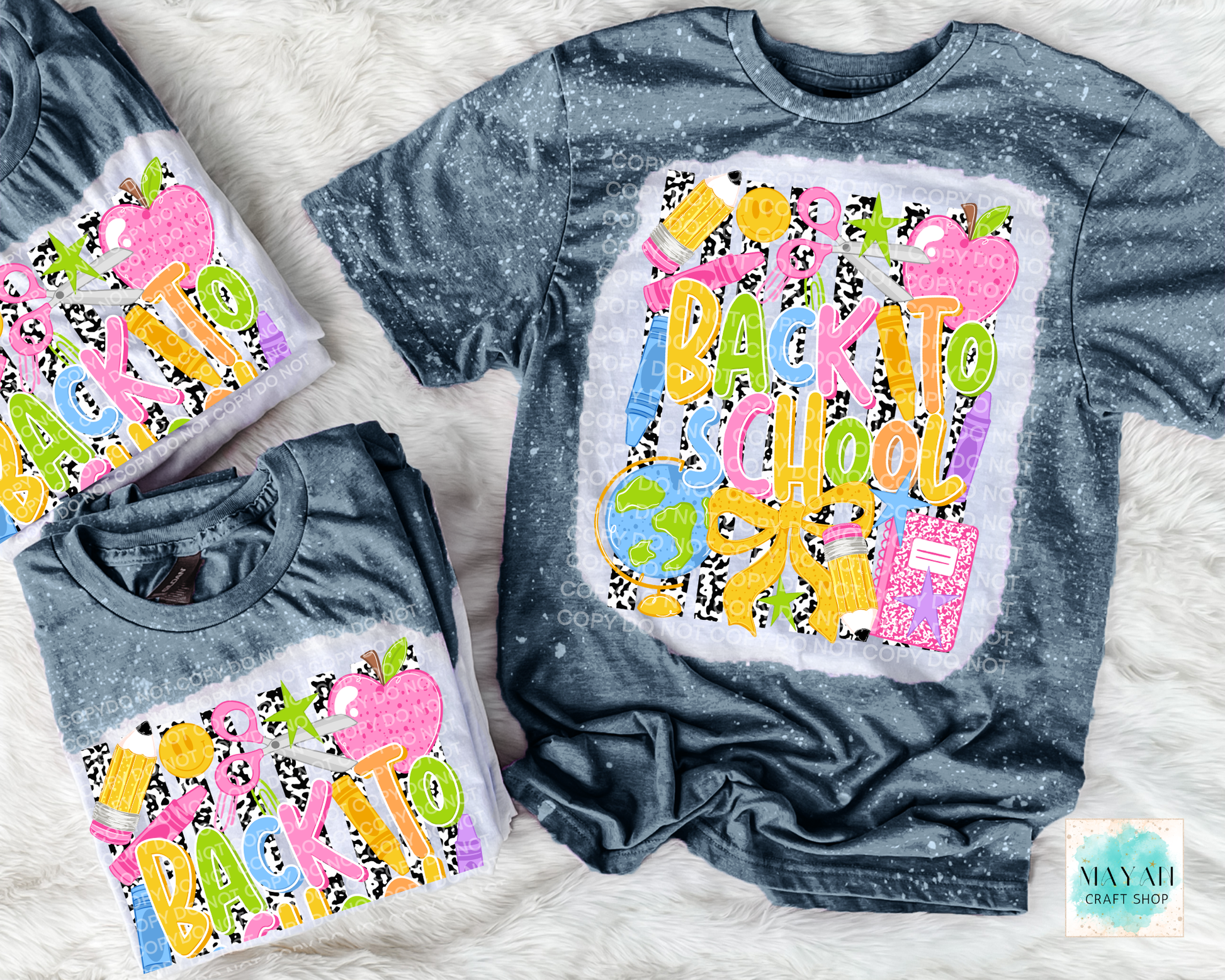 Three dark gray bleached t-shirts with a colorful 'Back to School' graphic, arranged on a white faux fur background. The graphic features school-themed illustrations like apples, a pencil, and a notebook in bright colors.