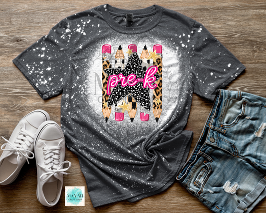 A dark gray splatter-print graphic t-shirt laid flat on a wooden surface, styled with white sneakers and distressed denim shorts. The t-shirt features a central graphic of pencils with leopard print patterns, a black star, and pink 'pre-k' text.