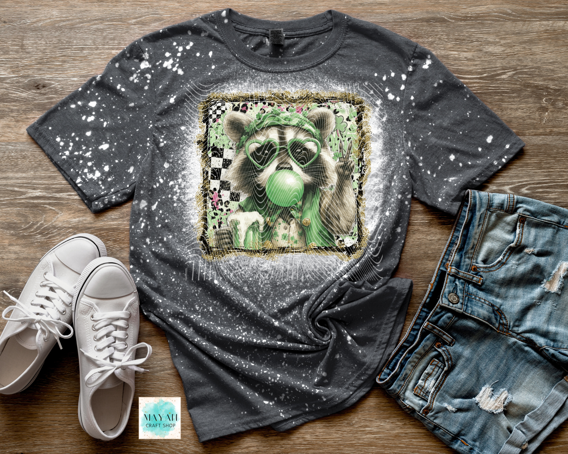 Gray t-shirt with a graphic design of a raccoon wearing a green hat, paired with white sneakers and blue jeans on a wooden surface.